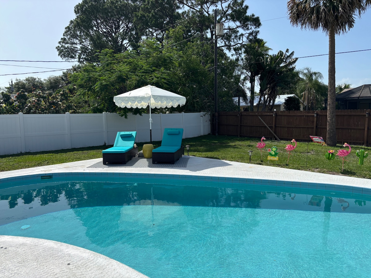 A serene backyard pool area is featured, with a clear blue pool surrounded by lush greenery. Two teal lounge chairs are positioned beneath a white canopy for shade. A fenced yard enhances privacy, while colorful lawn decorations add a cheerful touch.