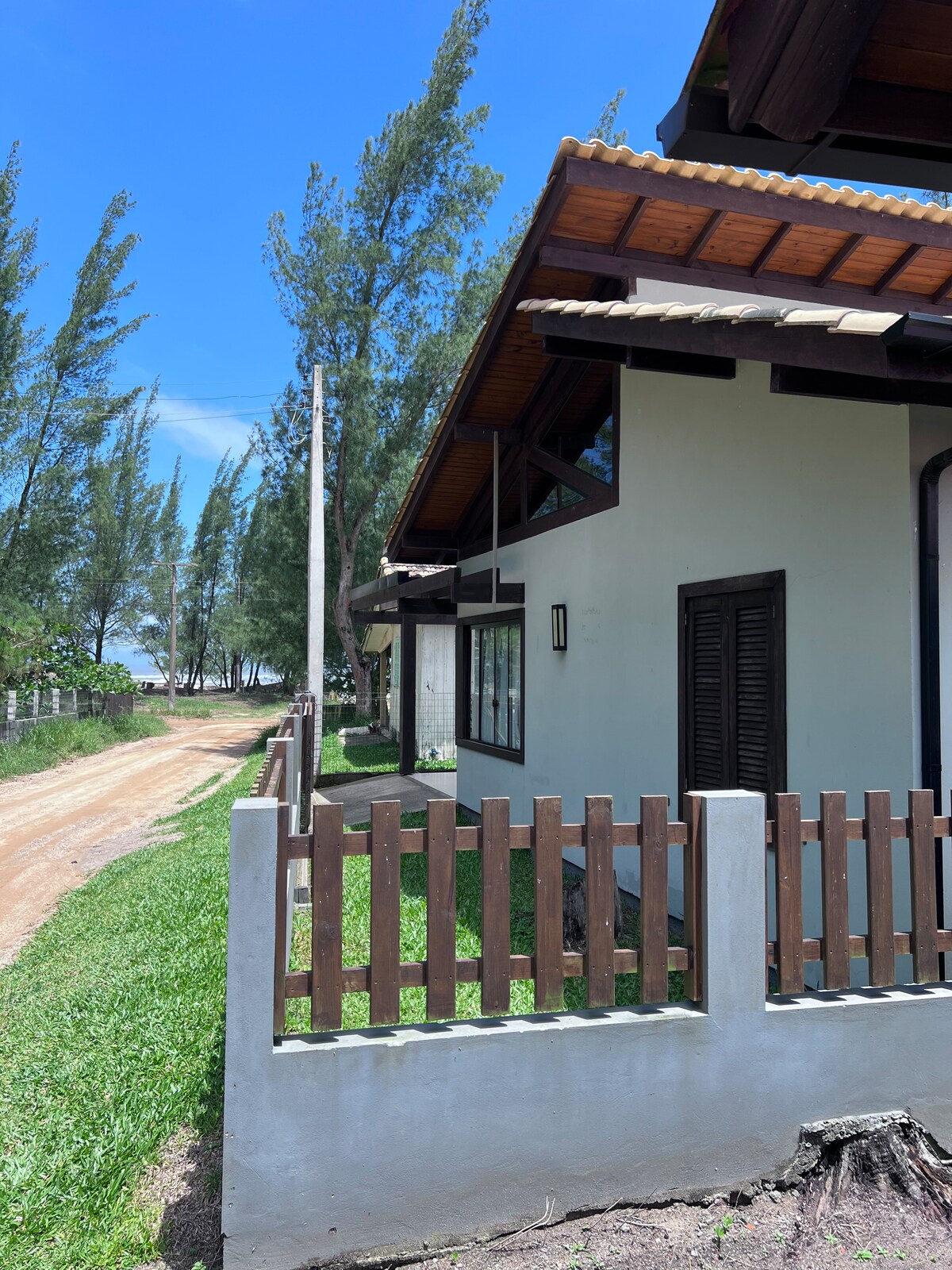 The exterior view of the house is presented, showcasing a well-maintained wooden fence surrounding the property. A pathway leads to the house, framed by green grass and tall trees, with the beach visible in the background under a clear blue sky.