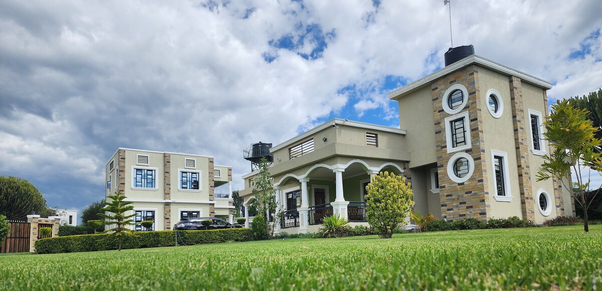 Two distinct buildings are set within a well-maintained lawn featuring manicured grass. One structure presents a modern design with circular windows, while the other showcases a more traditional façade, both complemented by a peaceful sky dotted with clouds.