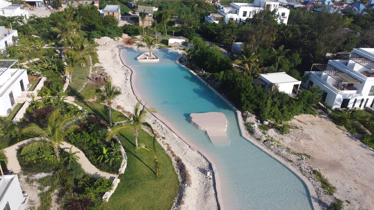 An aerial view of a serene landscape features a winding lagoon surrounded by lush greenery and landscaped areas. Sandy pathways meander along the water's edge, while contemporary villas are visible in the background, blending harmoniously with the natural surroundings.