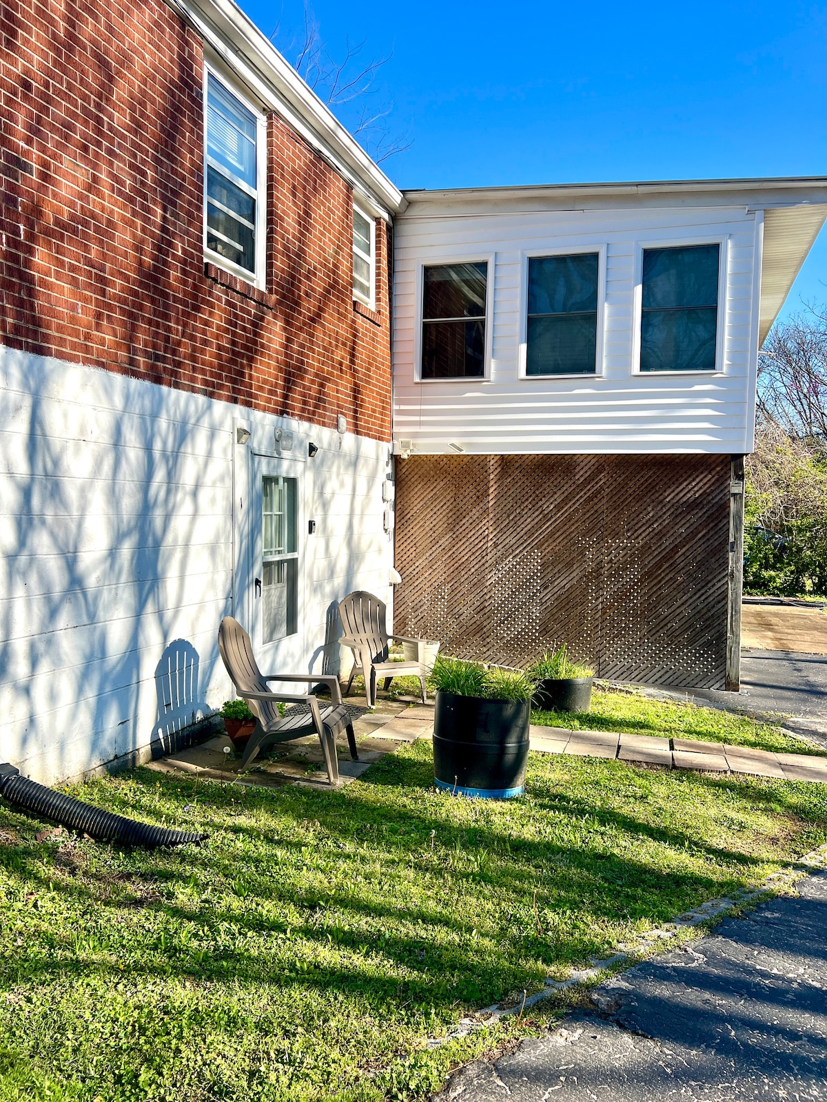 An exterior view of the property features a brick building adjacent to a white structure. Two wooden chairs are positioned on a grassy area, accompanied by a small potted plant. A barrel planter and stone path add a touch of greenery and accessibility to the entrance.