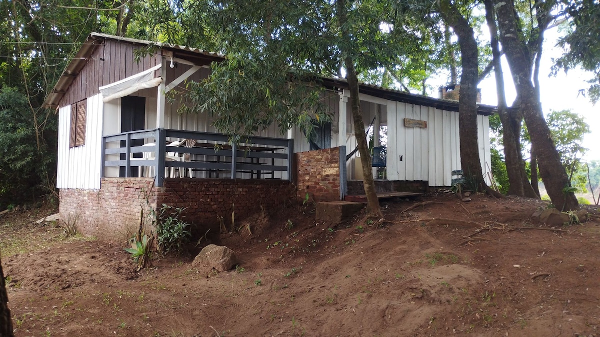 A simple house with white and dark wood siding is nestled among trees. A small porch extends from one side, leading to a pathway. The surrounding area features earthy tones and natural vegetation, creating a tranquil, nature-focused environment.