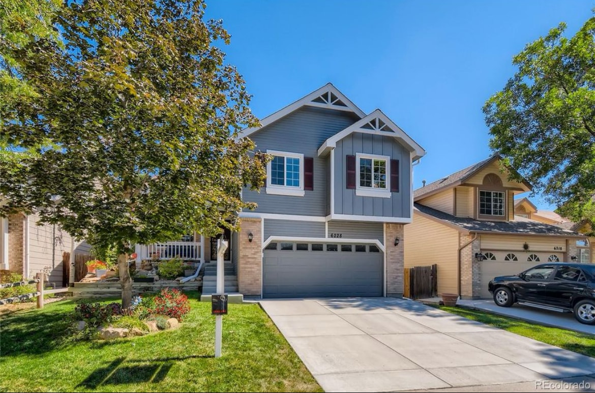 A two-story home is displayed with an inviting facade featuring gray siding and contrasting white trim. The well-maintained front yard is framed by trees and shrubs, while a concrete driveway leads to a two-car garage. Clear blue skies complete the pleasant exterior.