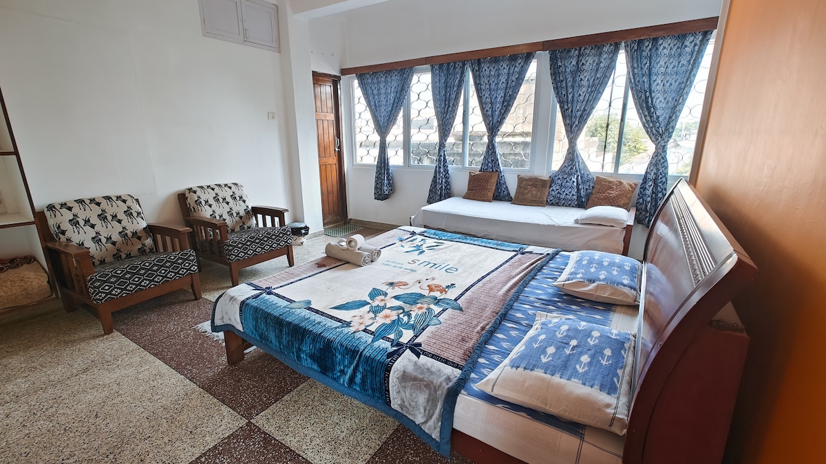 A spacious room features a large bed with a colorful blanket adorned with floral patterns. Two upholstered chairs are positioned nearby, with large windows allowing natural light to filter through the curtains. A sofa bed is also included, providing additional seating or sleeping space.