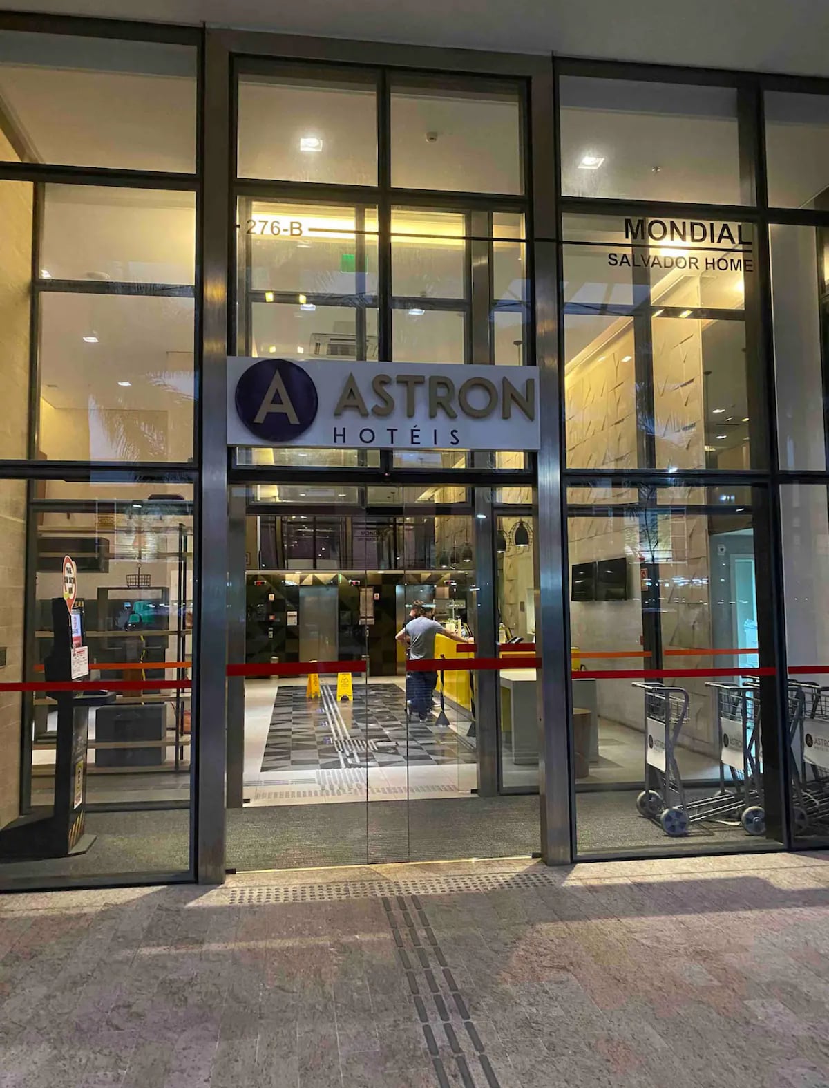 The entrance to Astron Hotéis is shown, featuring large glass doors with a prominent logo displayed above. A welcoming lobby area is visible inside, with a reception desk and various amenities. The space is well-lit, enhancing the modern design.