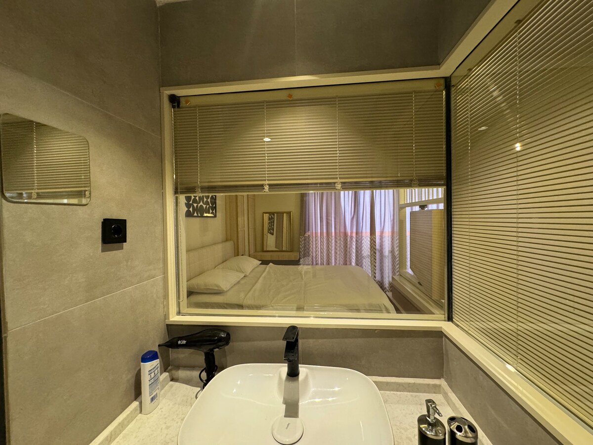 A modern bathroom is featured, with a spacious sink and a large mirror reflecting the soft lighting. A window with blinds overlooks a cozy sleeping area, where light drapes partially touch the bed. Sleek toiletries and a hairdryer are arranged on the counter.