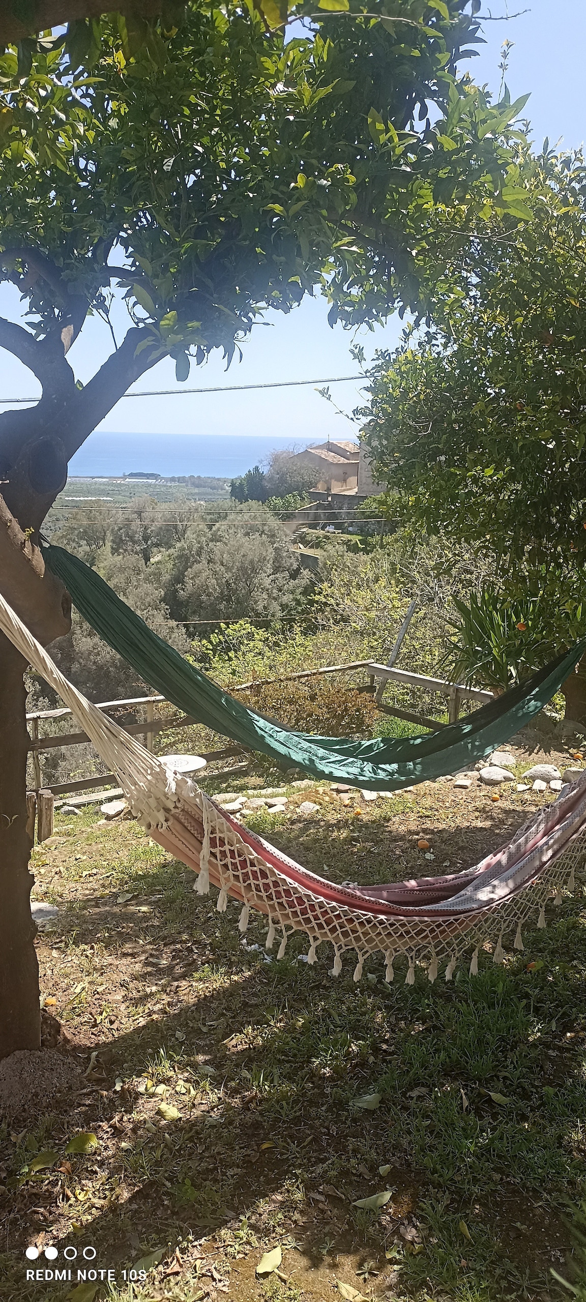 A spacious garden area features a hammock suspended between two trees, with greenery surrounding it. The view of the sea can be seen in the background, enhancing the outdoor experience. Sunlight filters through the branches, creating a serene atmosphere.