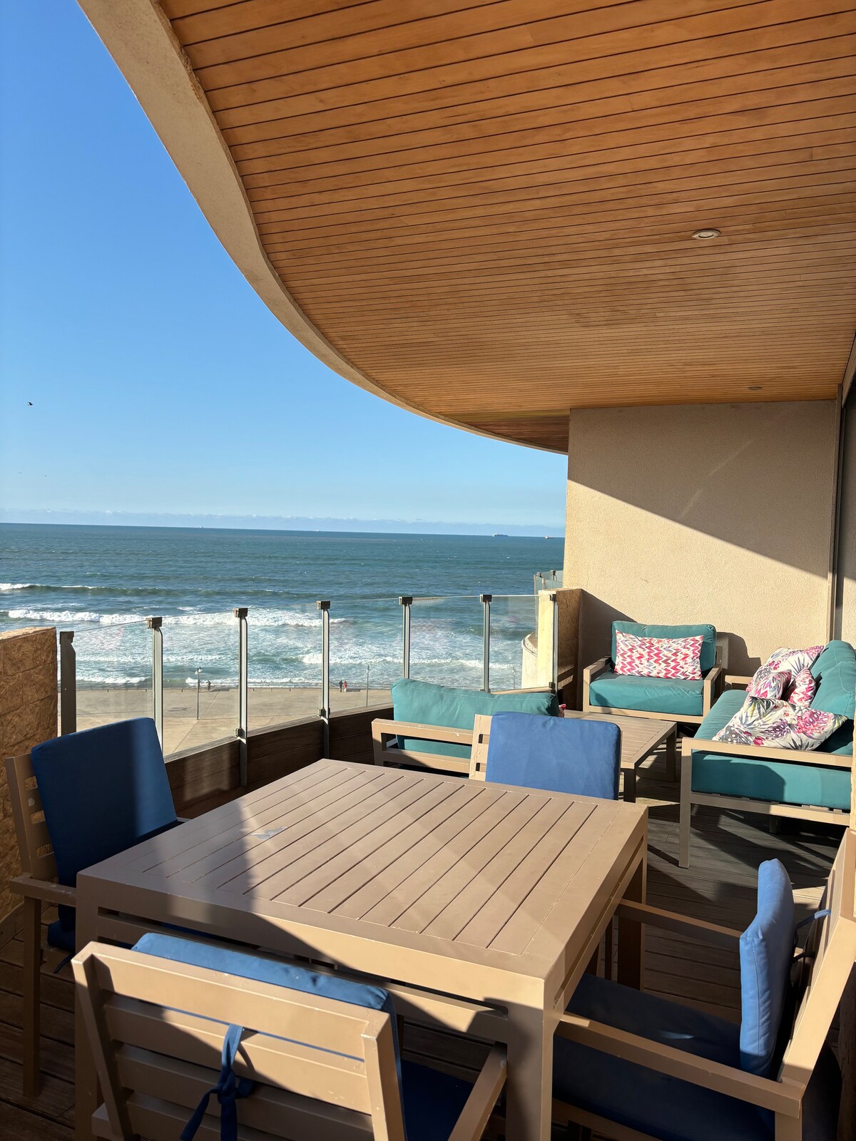 A private balcony features a dining table with several chairs, complemented by cozy seating. The ocean waves are visible in the background, while the sky above is clear and bright. Natural wood elements add to the serene atmosphere of this outdoor space.