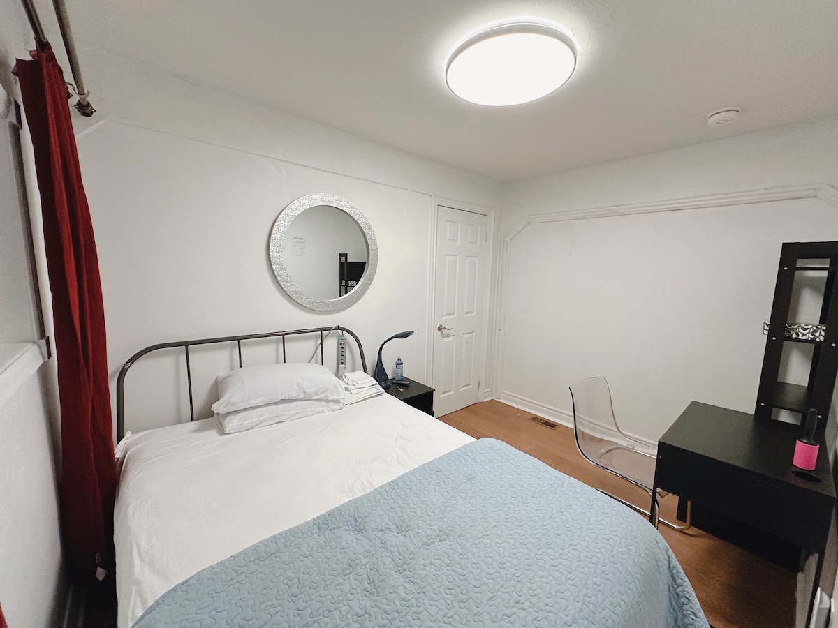 The room features a spacious bed with a soft, light blue coverlet. A circular mirror enhances the natural light, while a desk and transparent chair provide a work area. Red curtains add a pop of color against the neutral walls, and a closet door is visible.