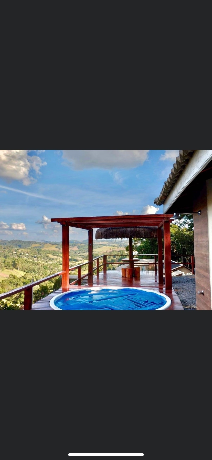 An outdoor terrace features a circular hot tub, offering expansive views of rolling hills under a clear sky. Wooden posts support a shaded area, while a small table and chairs provide a space for relaxation. Lush greenery surrounds the area, enhancing the natural setting.
