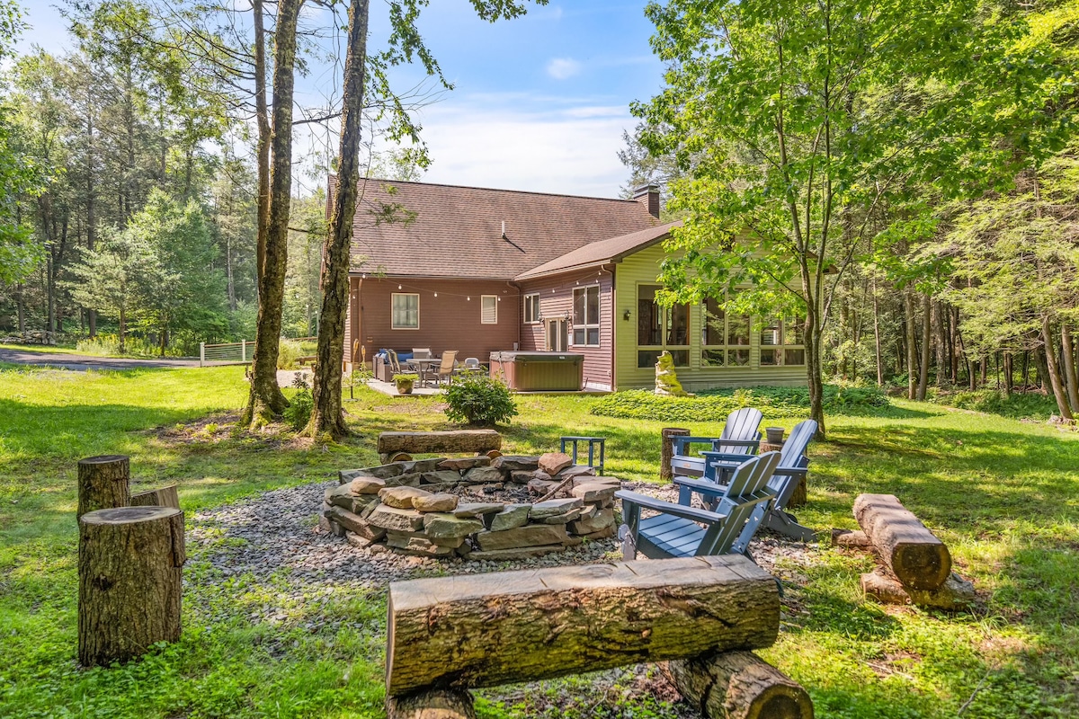 A serene outdoor area features a stone fire pit surrounded by log seating, nestled within a lush green landscape. The home is visible in the background, showcasing a mix of natural materials and large windows, inviting relaxation in a peaceful setting.