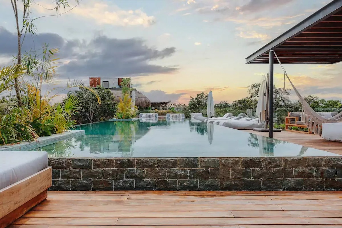 An inviting rooftop space features an infinity pool surrounded by wooden deck and sun loungers. Lush greenery sets the backdrop, while comfortable hammocks are suspended nearby. The sunset sky casts warm hues over the tranquil scene.