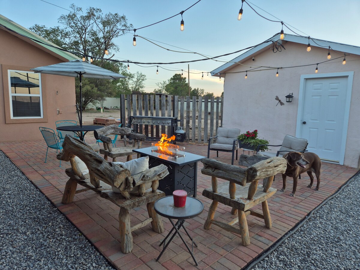 An outdoor gathering space features rustic log seating arranged around a central fire pit, which emits a warm glow. String lights are hung overhead, providing soft illumination. A table with a candle and an umbrella are also visible, creating a cozy atmosphere.