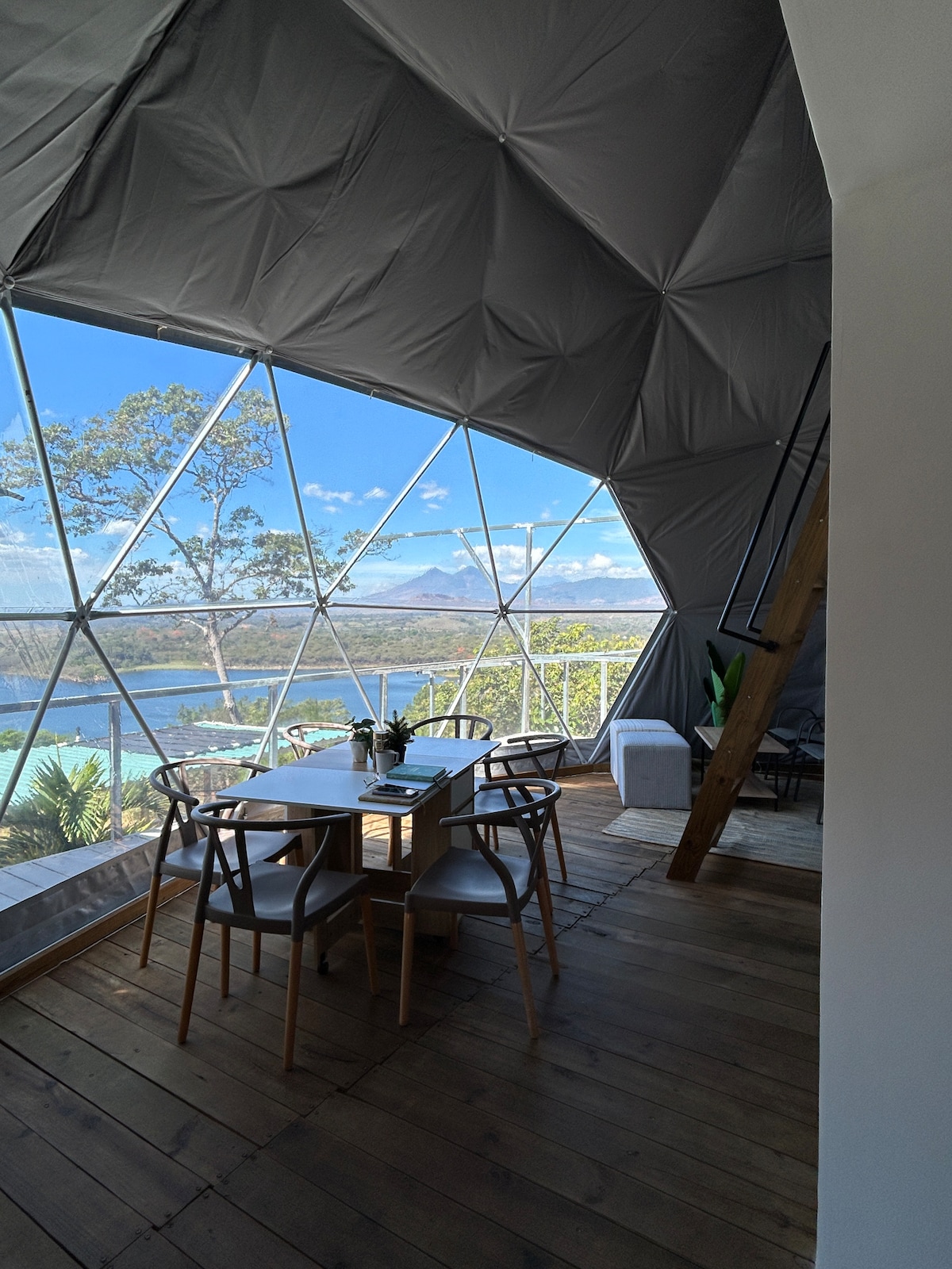 An interior view of the geodesic dome showcases a dining area with a simple table and chairs. Large windows provide a scenic view of the surrounding landscape, including a lagoon and distant volcanoes. Natural light enhances the wooden flooring and minimalist decor.