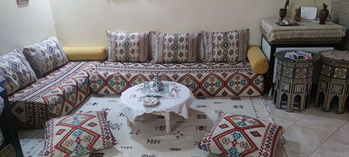 A cozy seating area is presented, featuring a low table surrounded by traditional cushions and patterned floor pillows. The walls display soft beige tones, while a richly patterned tapestry decorates the floor. Decorative pieces are placed on a side table, enhancing the inviting atmosphere.