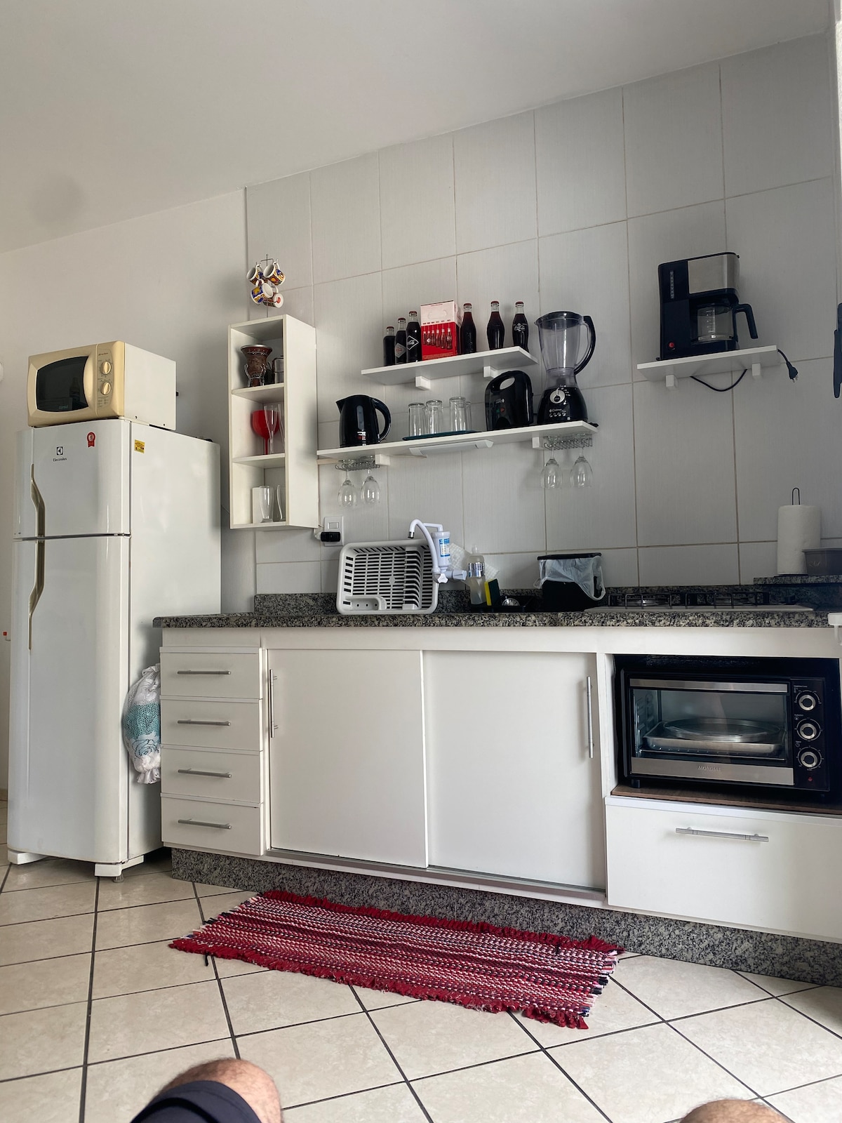 A modern kitchen is presented, featuring white cabinetry and granite countertops. Kitchen appliances such as a refrigerator, coffee maker, and microwave are visible. Glassware and utensils are neatly arranged on shelves, while a small woven rug adds a touch of warmth to the tiled floor.