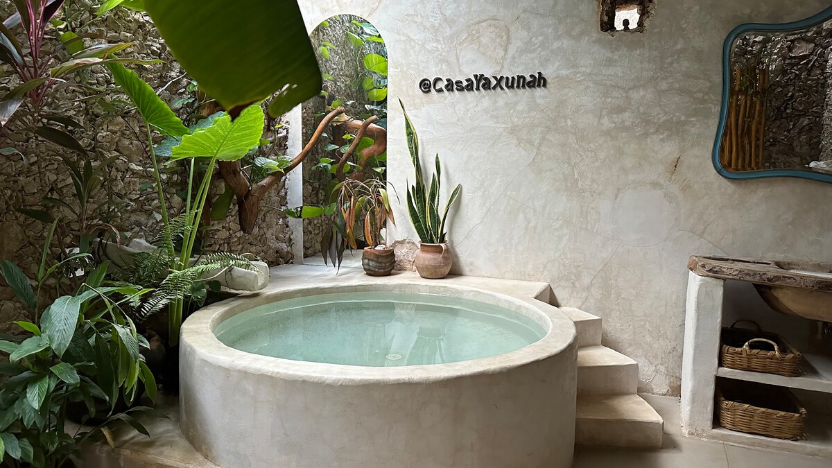 A sunlit space features a circular stone tub surrounded by lush tropical plants and textured walls. Natural light filters in through an archway, highlighting the calming tones of the polished surfaces. Decorative pottery and greenery accent the serene atmosphere.