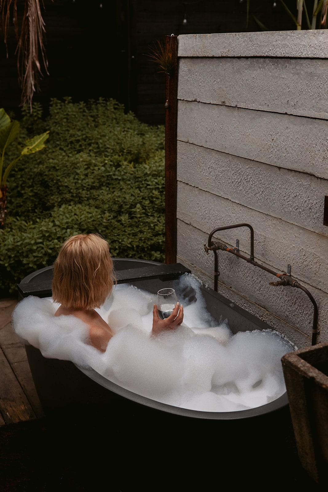 A soaking tub is positioned outdoors, surrounded by lush greenery. Bubbles fill the tub, while a glass sits comfortably in the occupant's hand. The wooden deck is visible, enhancing the natural ambiance of the setting.