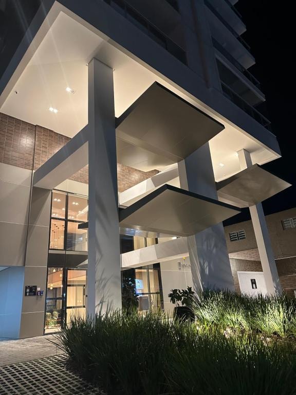 The exterior of a modern building is illuminated at night, highlighting geometric architectural elements. Clean lines and large glass windows create a contemporary appearance. Lush green plants and landscaping are visible at the entrance, adding a touch of nature to the urban environment.
