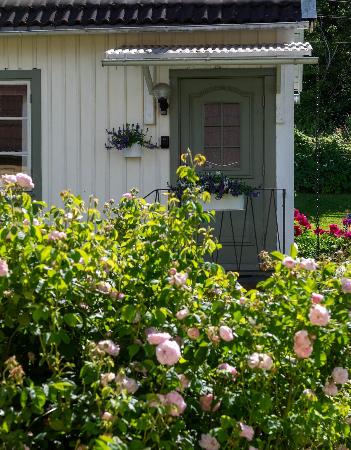 The entrance of a charming guest cottage is framed by lush green foliage and blooming pink roses. A green door features a decorative window and is accented by a small planter. The cottage background displays a well-maintained garden, enhancing the inviting appearance.