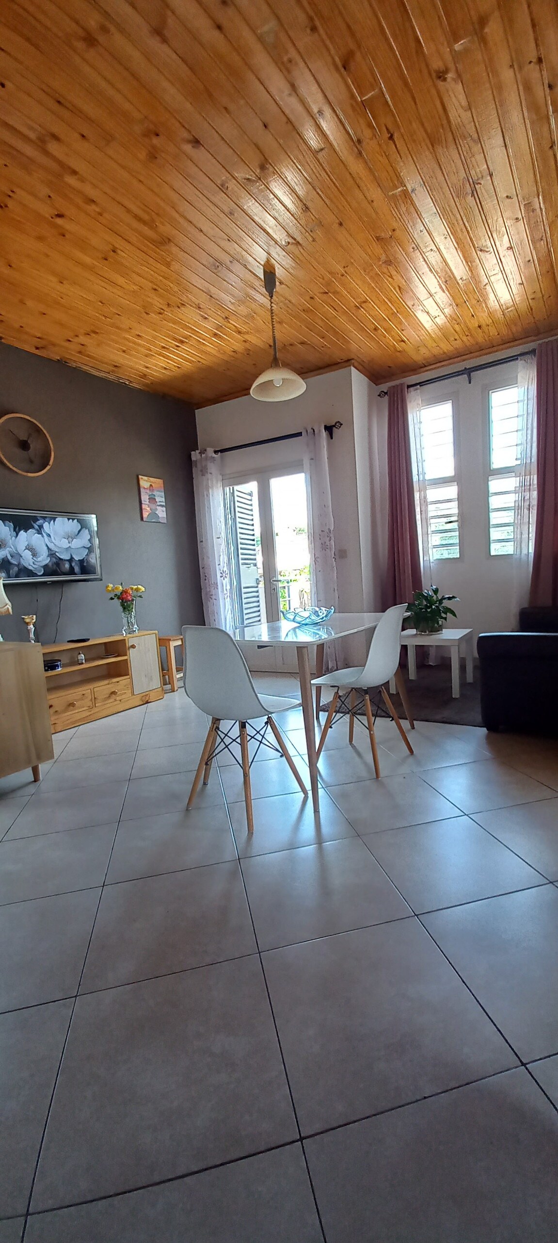 A bright living area features a wooden ceiling, enhancing the space with natural warmth. A round dining table is surrounded by modern chairs. Light streams through window panels adorned with sheer curtains, and a comfortable seating area with a television is visible in the corner.