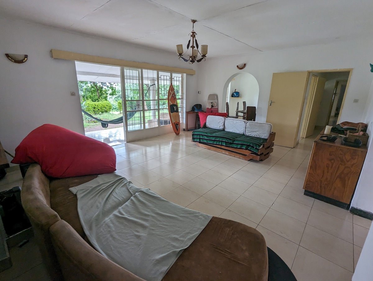 A spacious living area features a large brown couch with green and red cushions, set against a backdrop of white walls and tiled flooring. Natural light floods in through glass doors that open to a green outdoor space, creating a bright and airy environment.