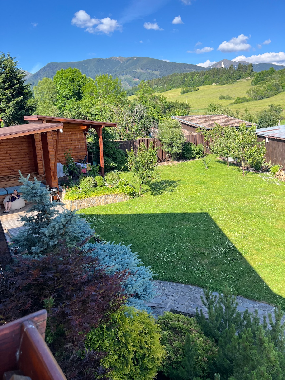 A spacious garden is visible, featuring green lawns and various trees. A wooden gazebo is seen among the lush vegetation, providing a shaded seating area. In the background, rolling hills and mountains create a scenic backdrop under a clear blue sky.
