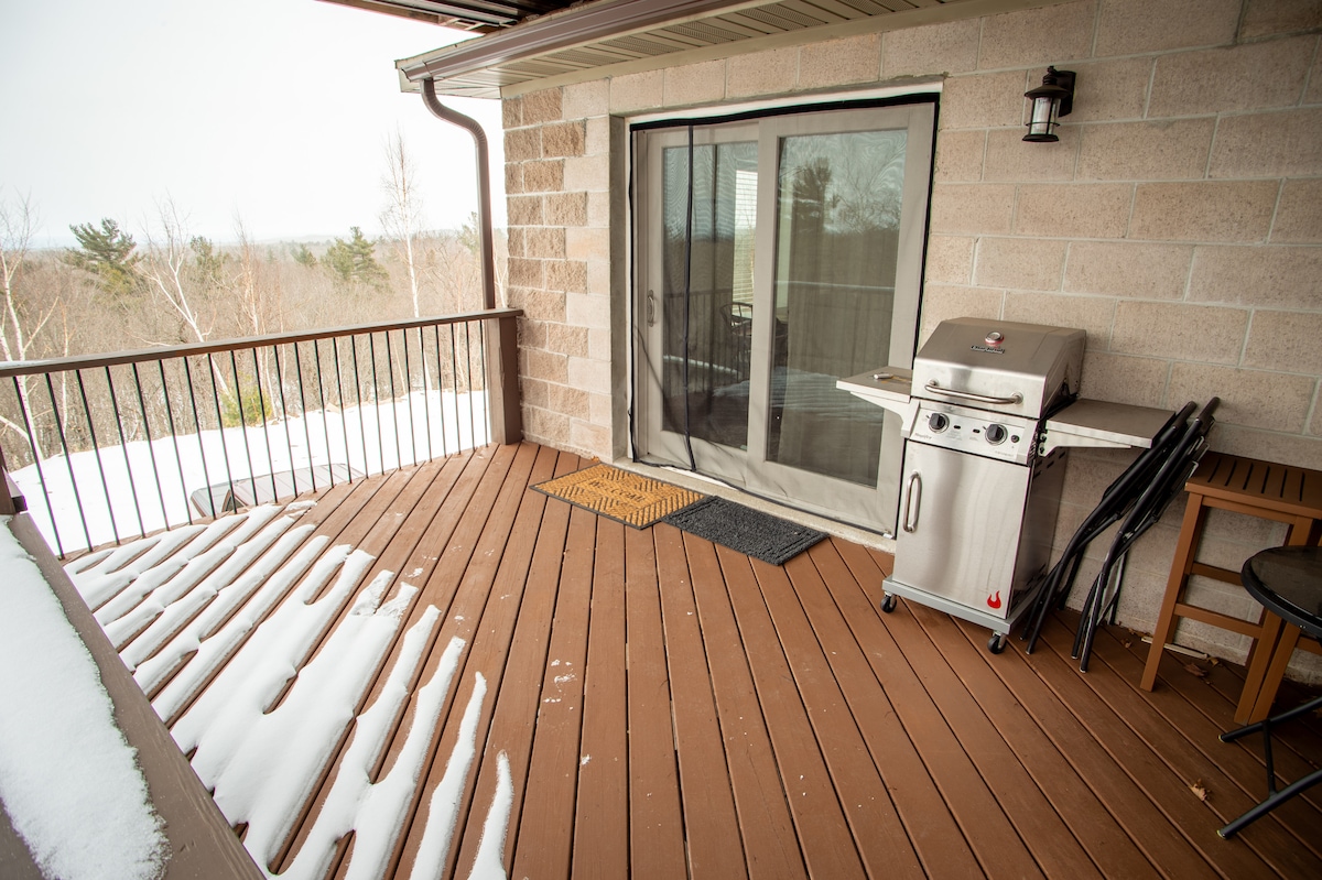 A spacious outdoor deck features wooden flooring partially covered with snow. Sliding glass doors open onto the deck, allowing access to the interior. A propane grill is positioned on one side, accompanied by a small table and chair set near the railing overlooking the natural surroundings.