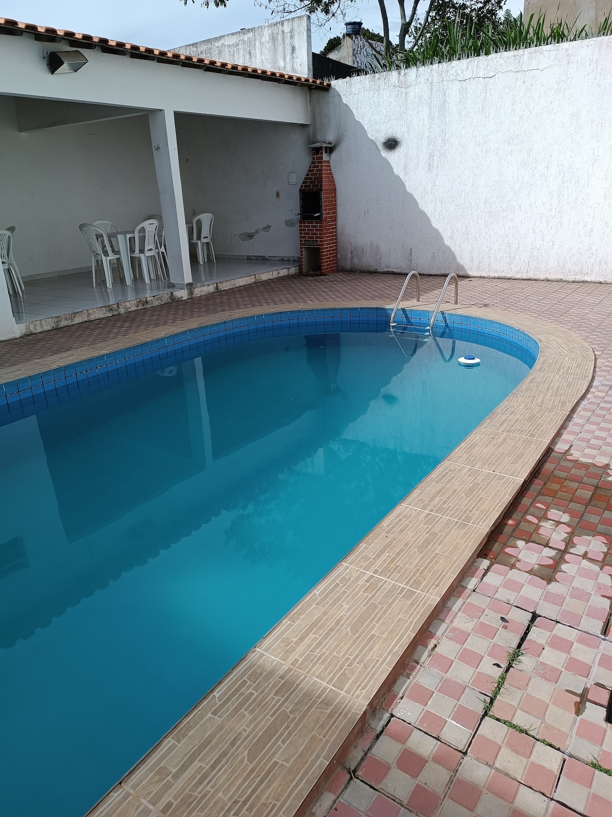 A clear swimming pool is depicted, featuring a light blue surface and a shallow end with two ladders. Surrounding the pool, a tiled patio with a patterned design is visible, as well as a shaded area with several white chairs and a barbecue grill.