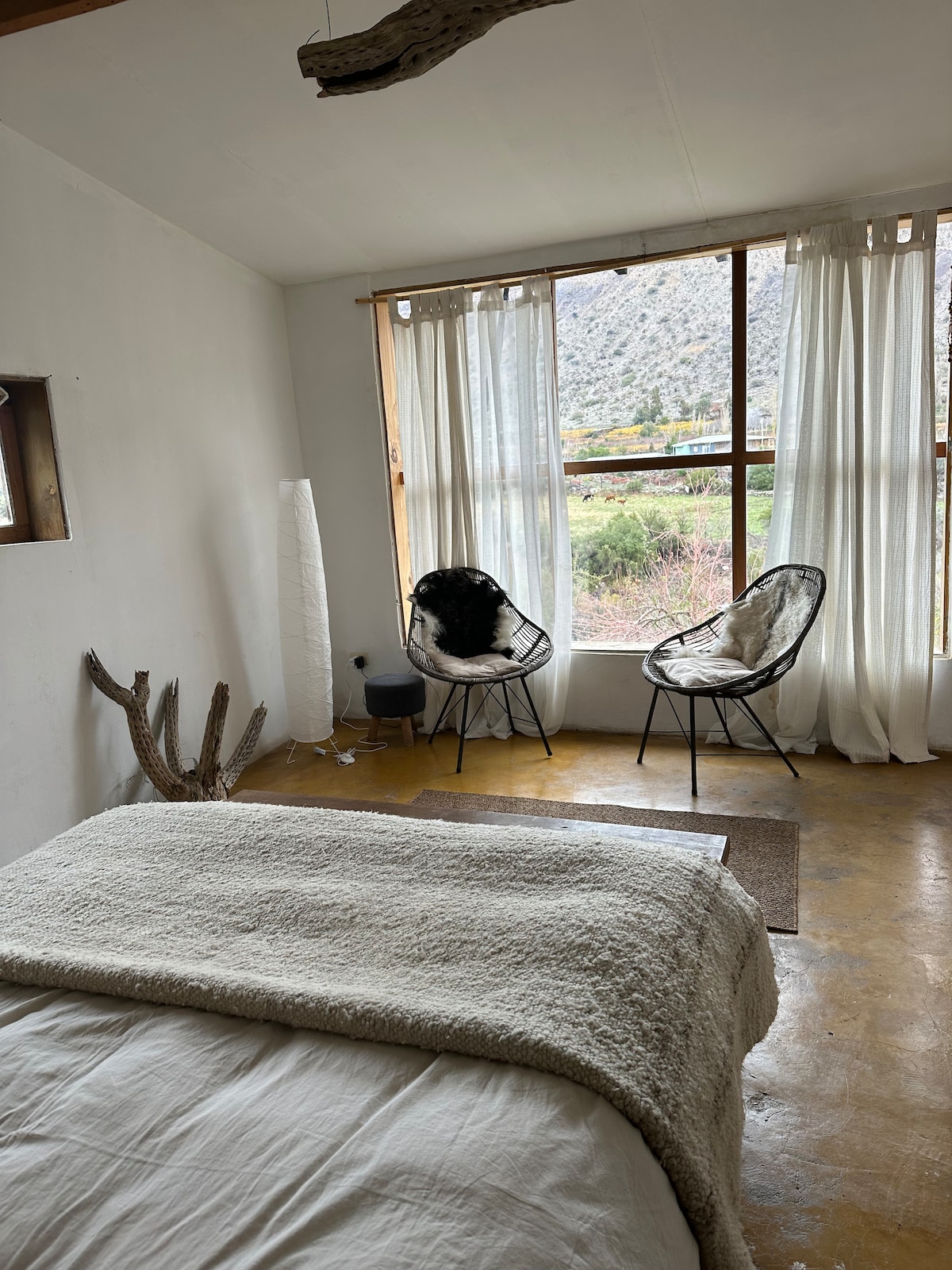 A bright bedroom features a spacious bed covered with a light blanket. Two comfortable chairs are positioned near a large window, providing views of the surrounding landscape. Sheer curtains allow natural light to filter in, creating an airy atmosphere.