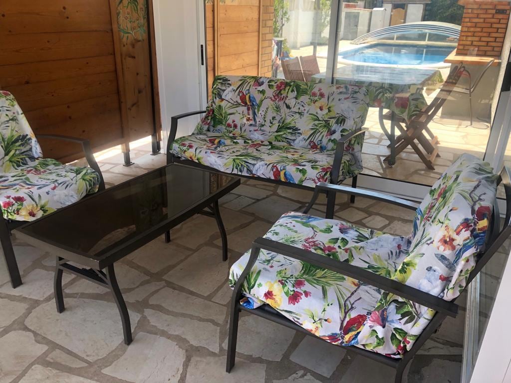 A sunlit sitting area features four chairs and a coffee table. The chairs are adorned with floral upholstery, providing a vibrant contrast to the stone floor. In the background, a swimming pool is visible through glass doors, enhancing the inviting ambiance of the space.