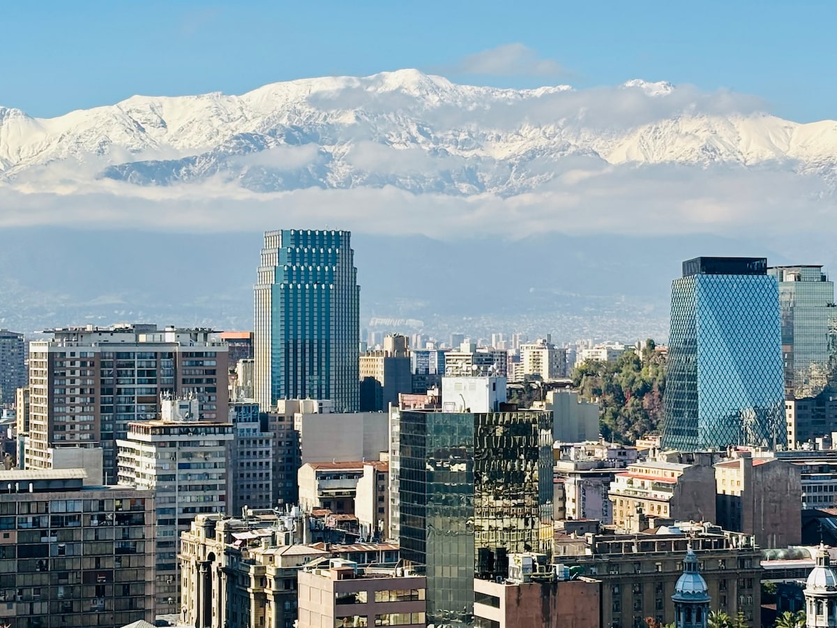 A panoramic view of Santiago, showcasing a mix of modern architecture and historic buildings. The backdrop features snow-capped mountains under a clear blue sky, with urban structures of varying heights and designs filling the foreground.