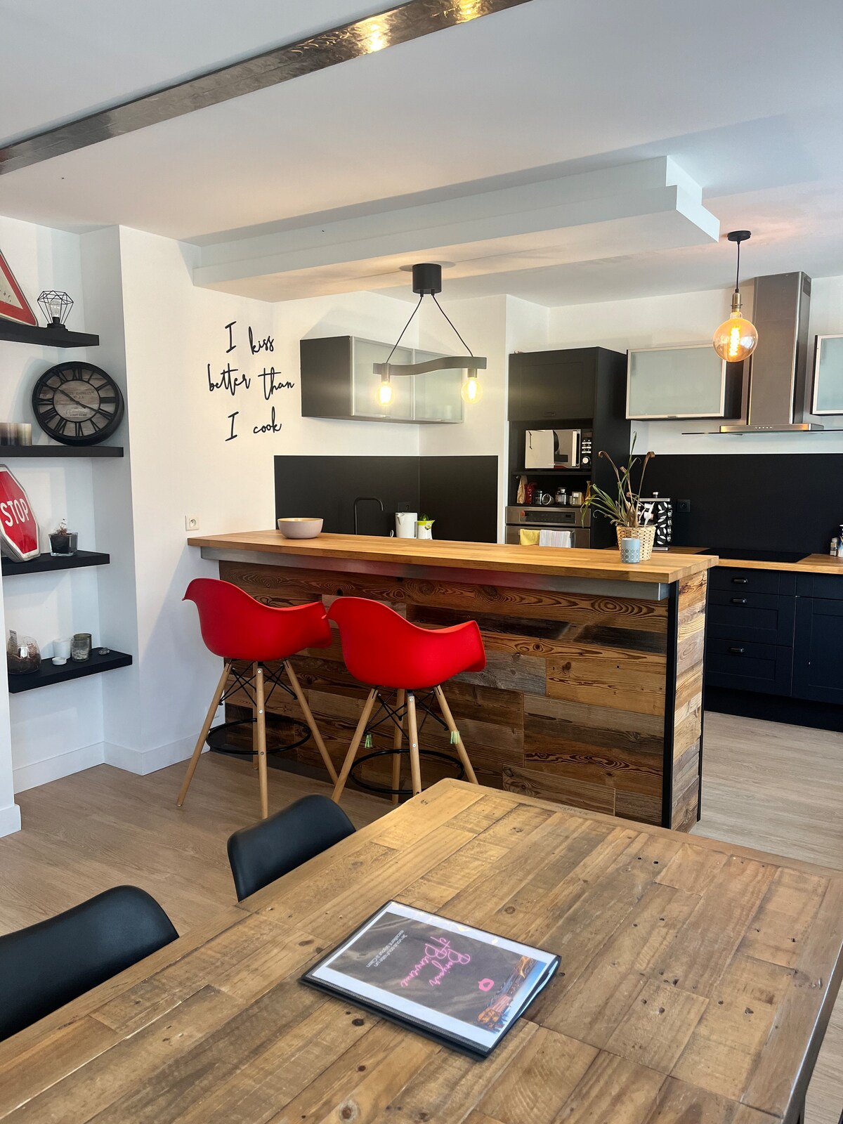 A modern kitchen and dining area are featured, with wooden accents and a bar island. High red stools are positioned at the bar, while a wooden dining table is set with dark chairs. Decorative items and plants are visible, enhancing the functional design.