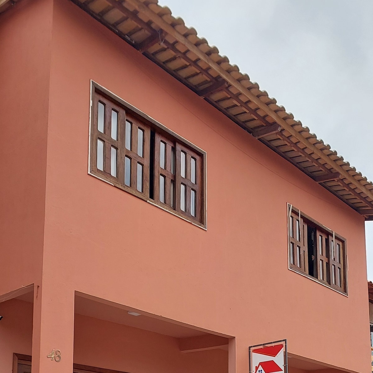 The exterior of a two-story structure is presented, featuring light peach-colored walls. Multiple wooden-framed windows are visible on the upper level, allowing natural light to enter. A tiled roof completes the design, with a prominent sign partially seen at the bottom.