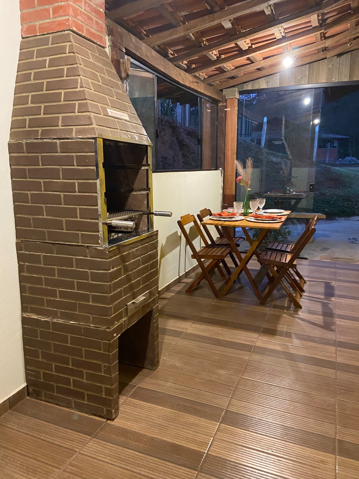 An outdoor barbecue area features a brick grill and a wooden table set with utensils. Two folding chairs are positioned around the table, which is placed under a wooden roof with warm lighting, creating a cozy atmosphere. A grassy area is visible beyond the patio.