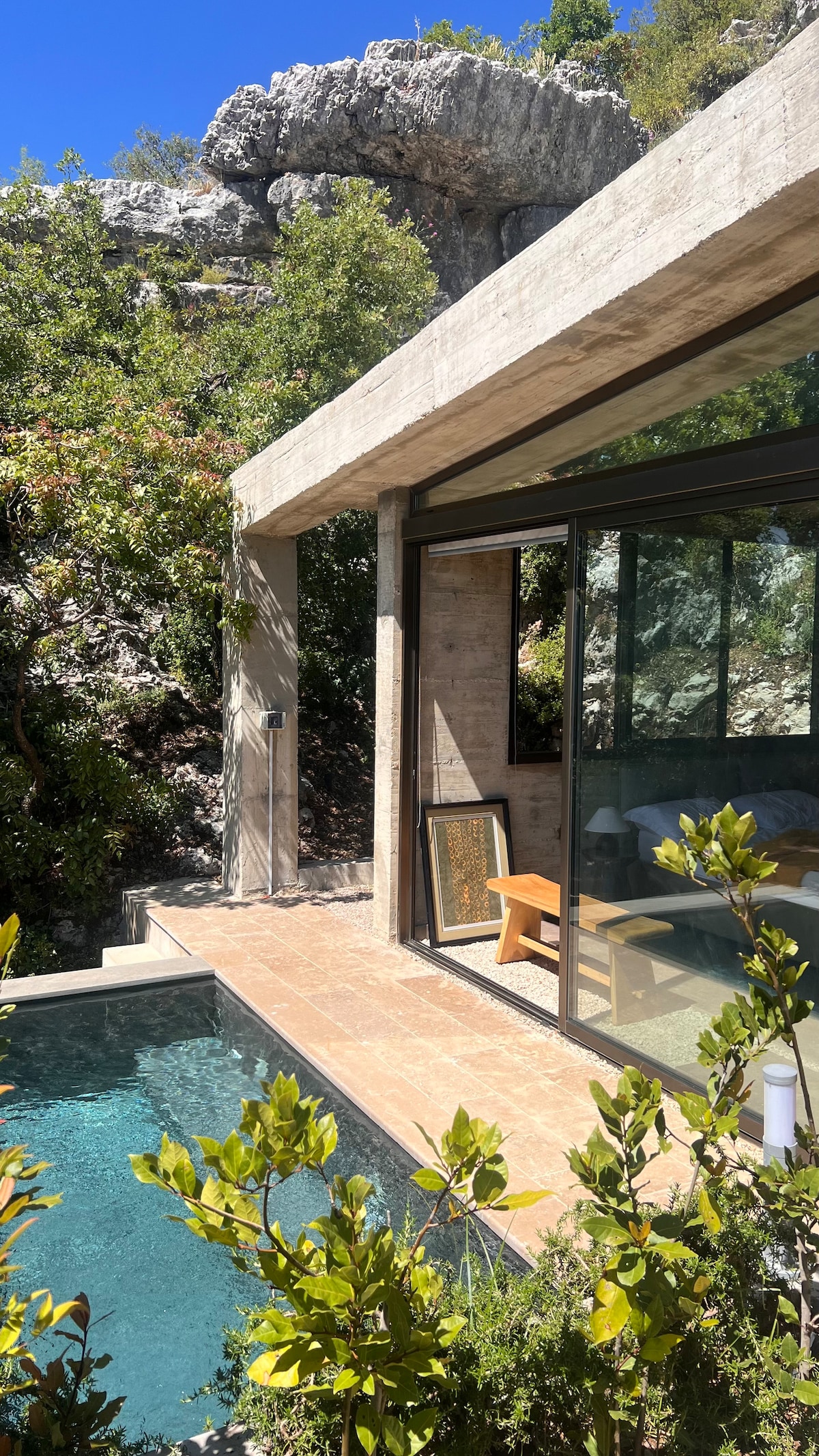 A modern villa is framed by lush greenery and rocky terrain. Large glass panels invite natural light into the living area, and a small pool reflects the surrounding foliage. A wooden chair is positioned on the patio, providing a view of the tranquil landscape.