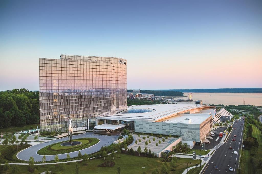 A modern casino resort stands prominently against the evening sky. The building features a glass exterior reflecting the sunset, while landscaped gardens with sculptures adorn the front entrance. The surrounding area includes a roadway and views of water in the background.