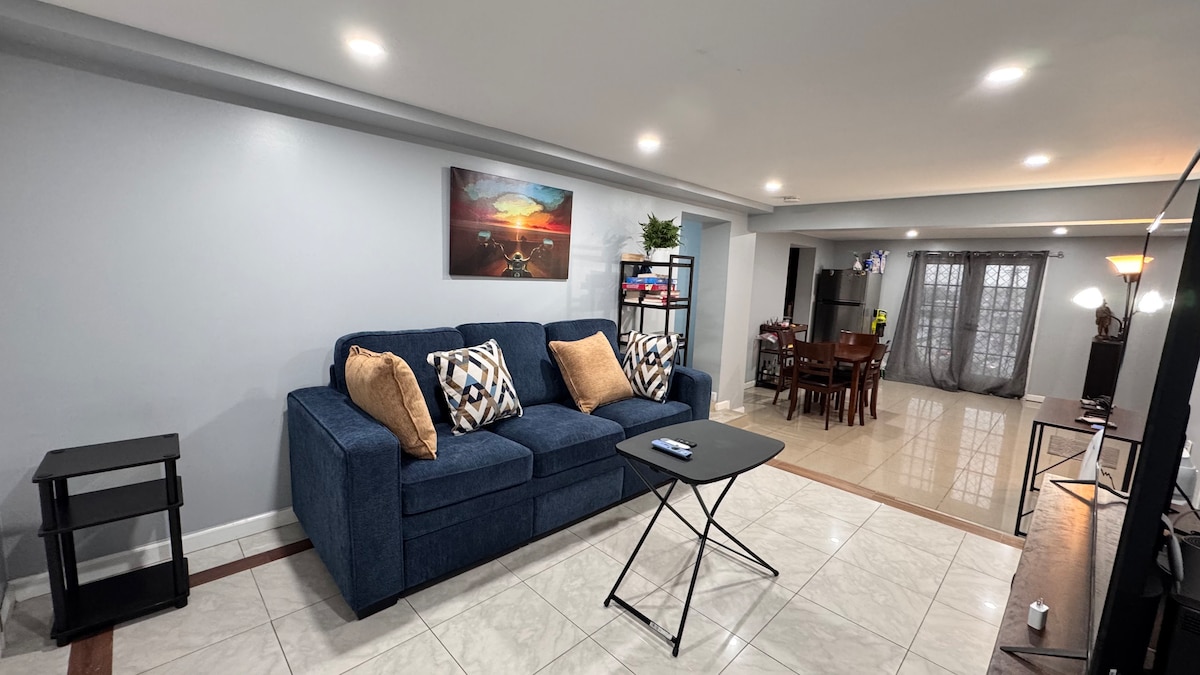 A spacious living area features a blue couch adorned with patterned throw pillows, positioned near a small folding table. Natural light filters through sheer curtains, illuminating a dining space with a dark wood table and chairs, enhancing the open layout.