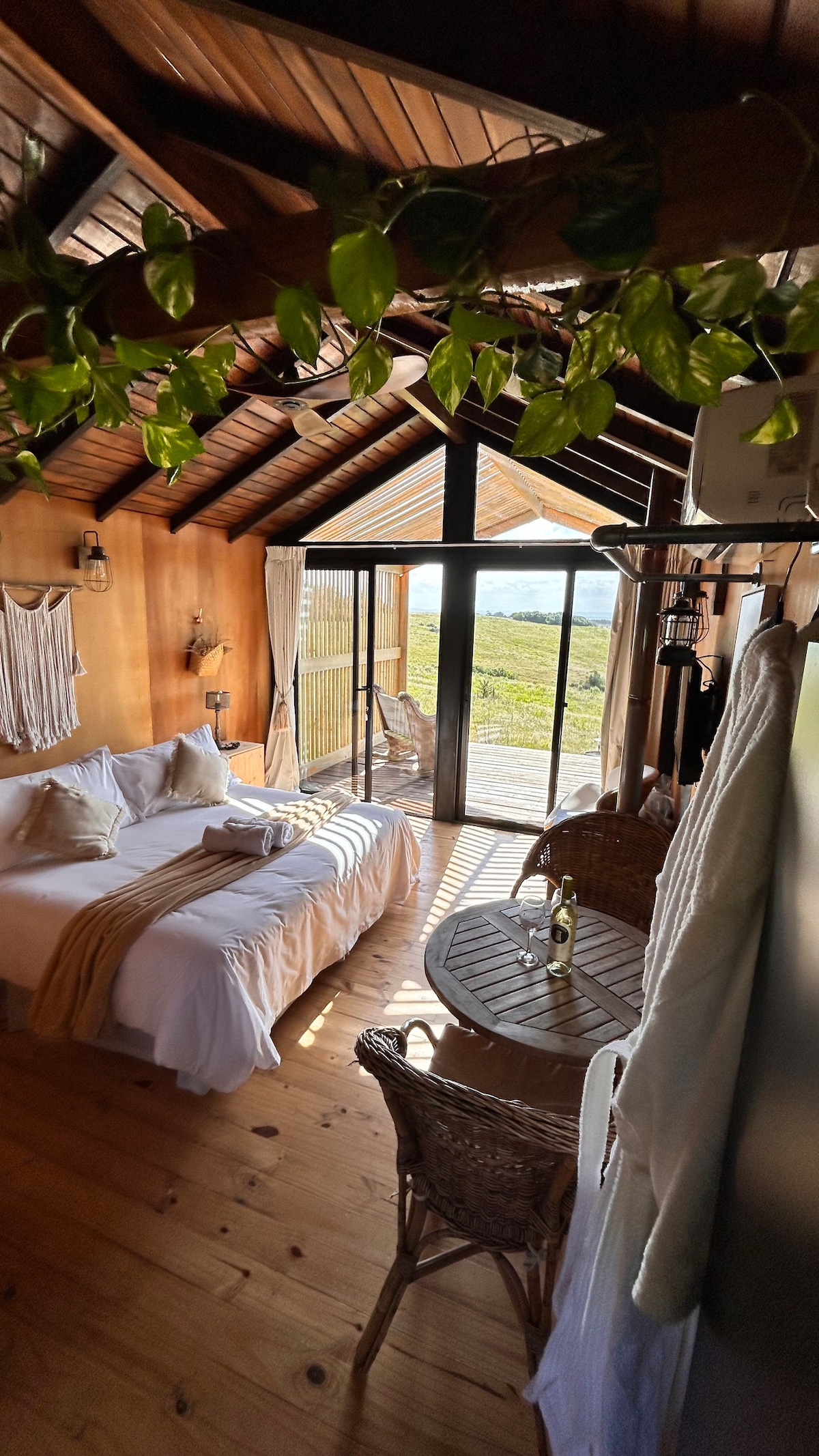 An airy room features wood-paneled walls and a high ceiling with exposed beams. A spacious bed adorned with soft linens rests beside a round table and two wicker chairs. Large glass doors open to a scenic view of nature, inviting the outdoors in.