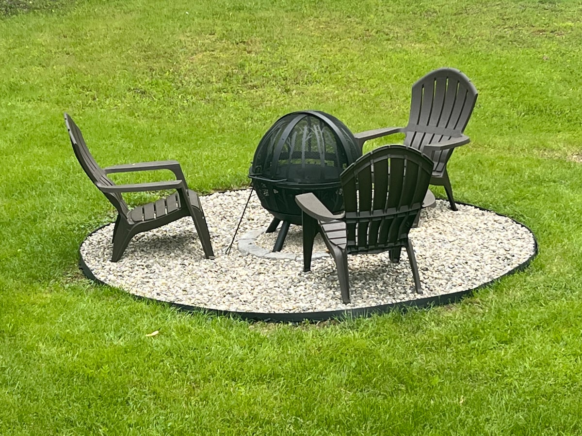 A circular gravel area is surrounded by a grassy landscape, featuring a black fire pit at its center. Four dark-colored adirondack chairs are positioned around the pit, providing a cozy seating arrangement for gatherings.