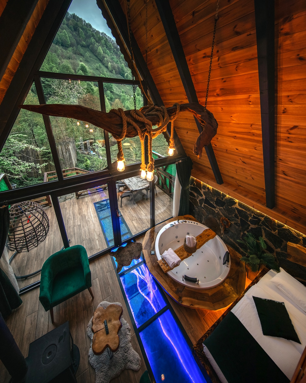 An open indoor space features a large jetted tub surrounded by natural wood accents. Large windows provide views of the surrounding greenery, while unique lighting fixtures hang from the ceiling. A cozy seating area is arranged with plush chairs and a wooden coffee table.