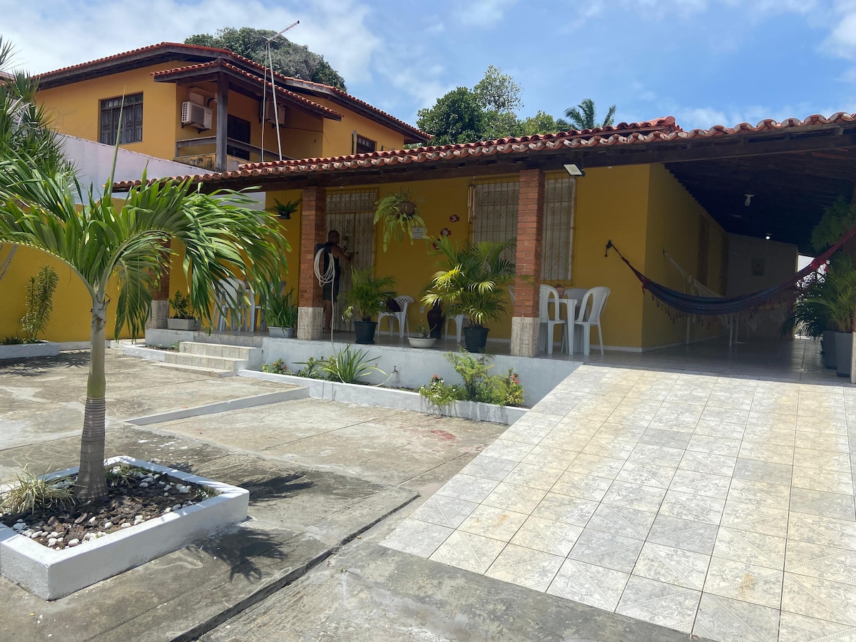 An inviting exterior view of the house is presented, featuring a spacious patio with white chairs and a hammock. Lush greenery surrounds the entrance, and a paved walkway leads to the entrance. The vibrant yellow facade contrasts with the tiled roof and blue sky.