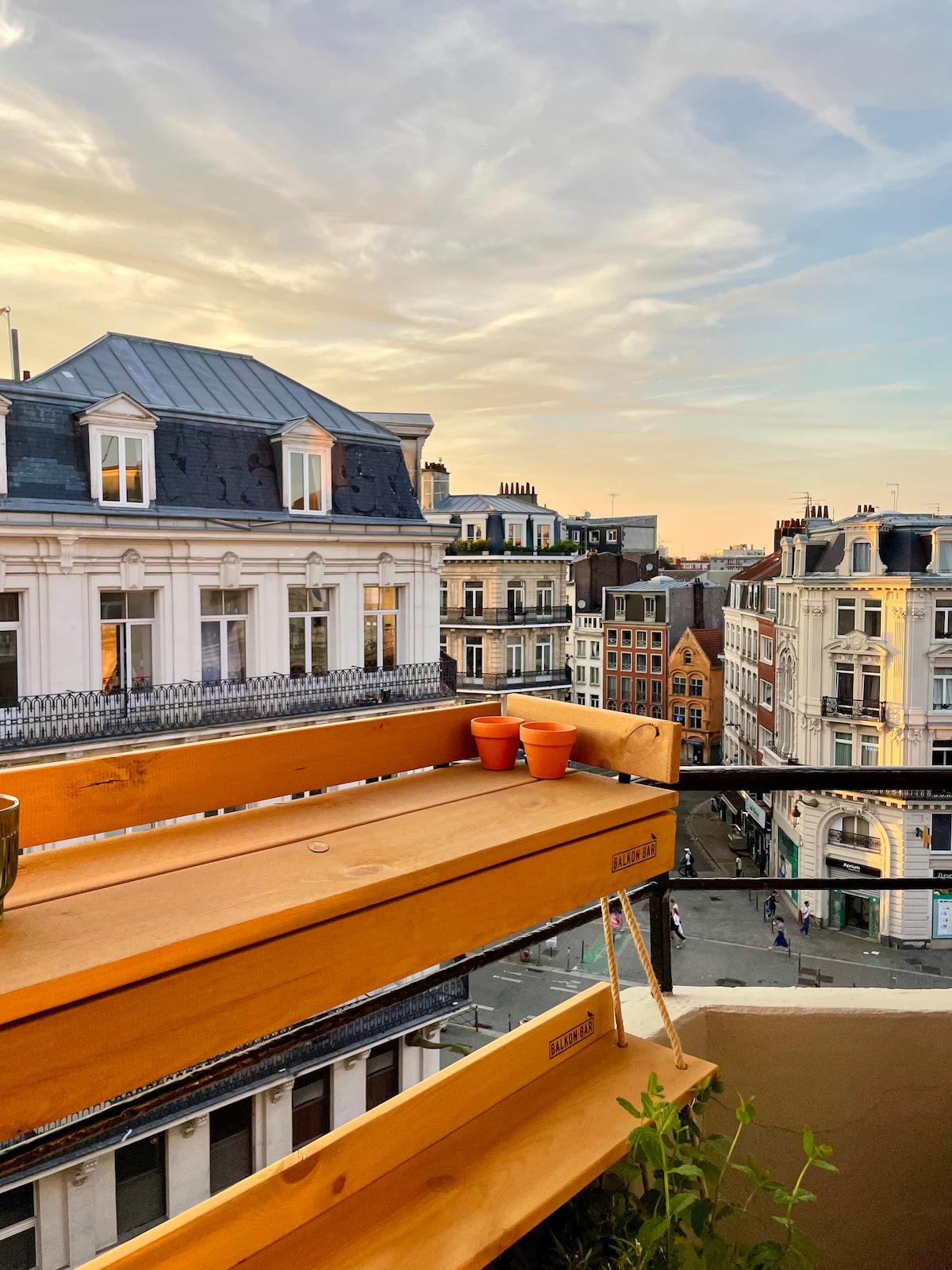 A wooden balcony railing is prominently displayed, adorned with small terracotta pots. In the background, rooftops of various buildings are visible, along with a vibrant sky at sunset, blending shades of orange and blue, creating an expansive view of the surrounding cityscape.