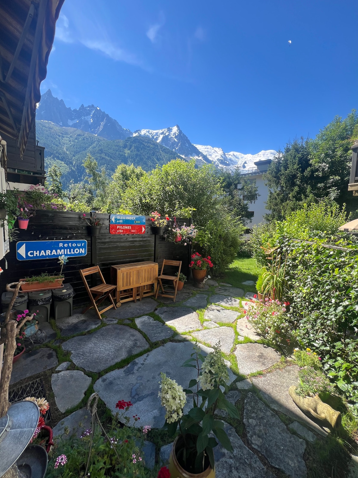 A private garden area is seen, featuring natural stone pathways and vibrant greenery. Wooden chairs sit beside colorful flower pots, while scenic views of Mont Blanc and surrounding mountains are visible in the background under a clear blue sky.