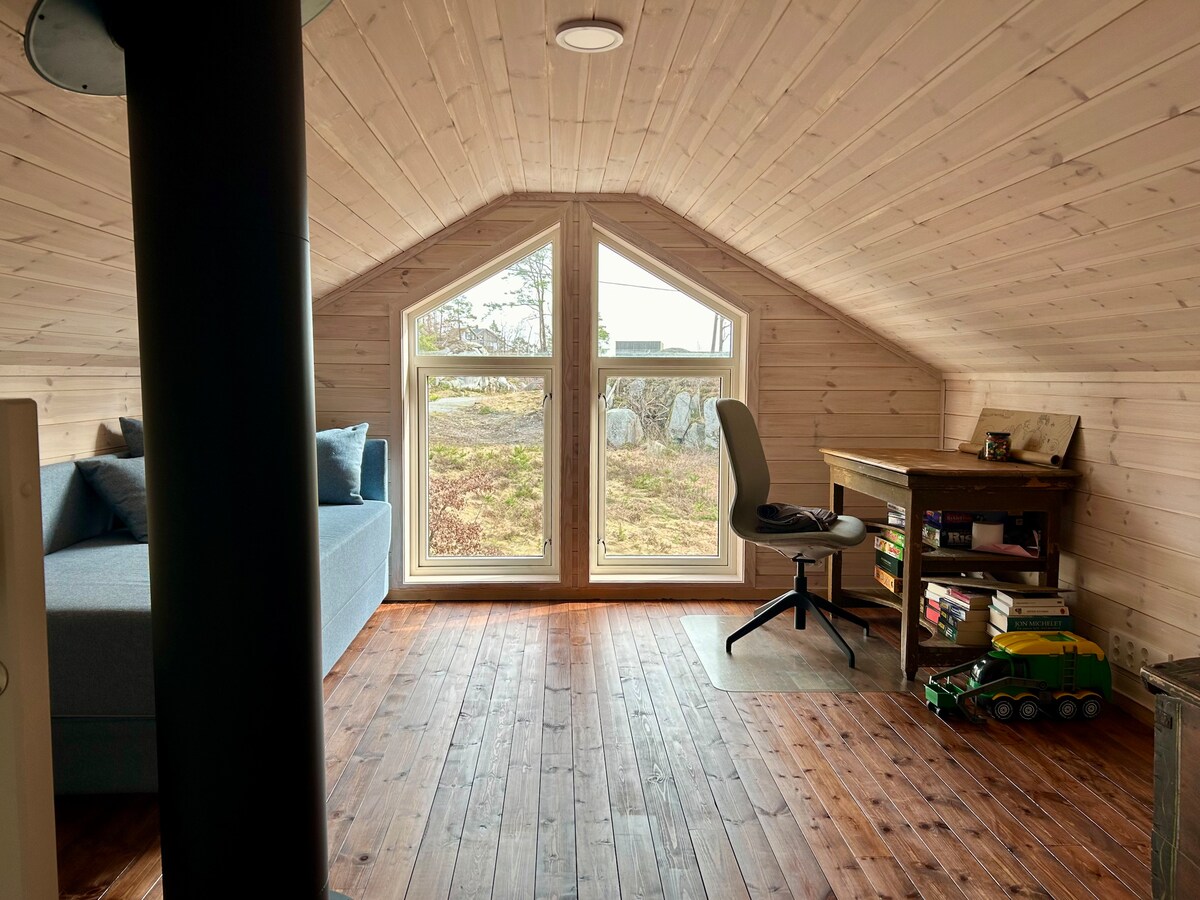 A loft area features a cozy seating arrangement with a sofa and a small wooden desk. Two large windows provide natural light and views of the outdoor scenery. Wooden flooring complements the wooden walls, creating a warm, inviting space.