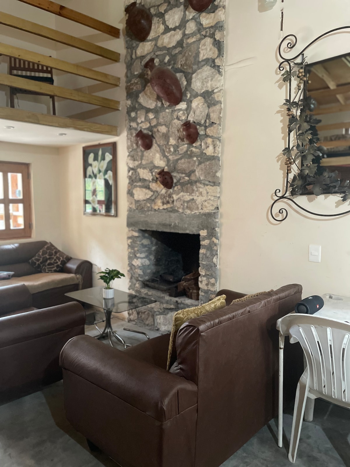 A cozy living area is highlighted by a stone fireplace, with artistic decorations positioned above it. Two brown armchairs are arranged around a glass coffee table. A small dining table and chair can be seen in the background, alongside large windows that allow for natural light.