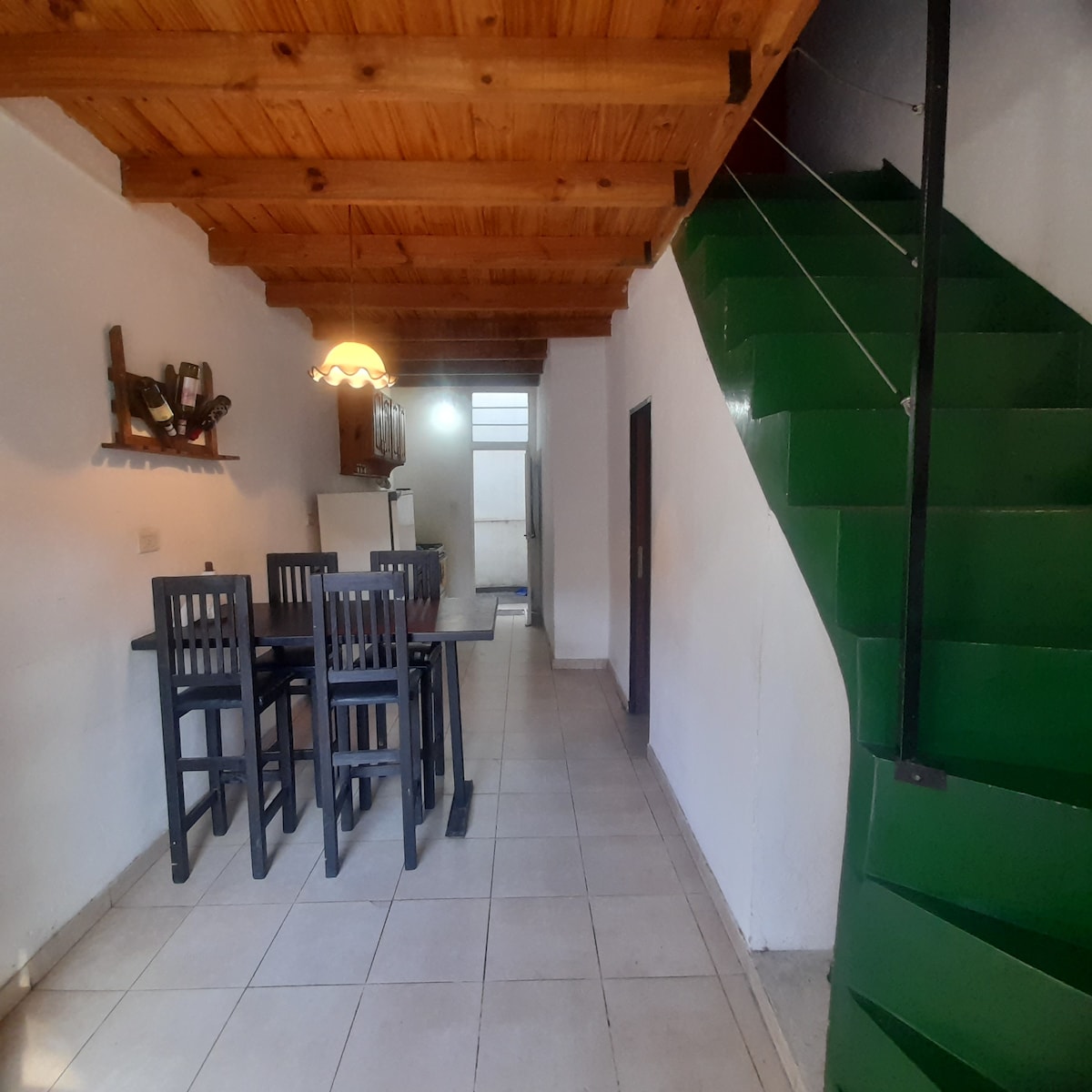 A spacious dining area is complemented by a high wooden ceiling and a large window allowing natural light. A wooden dining table with tall chairs is positioned near the entrance. A distinctive green staircase leads to the upper level, adding a modern touch to the design.
