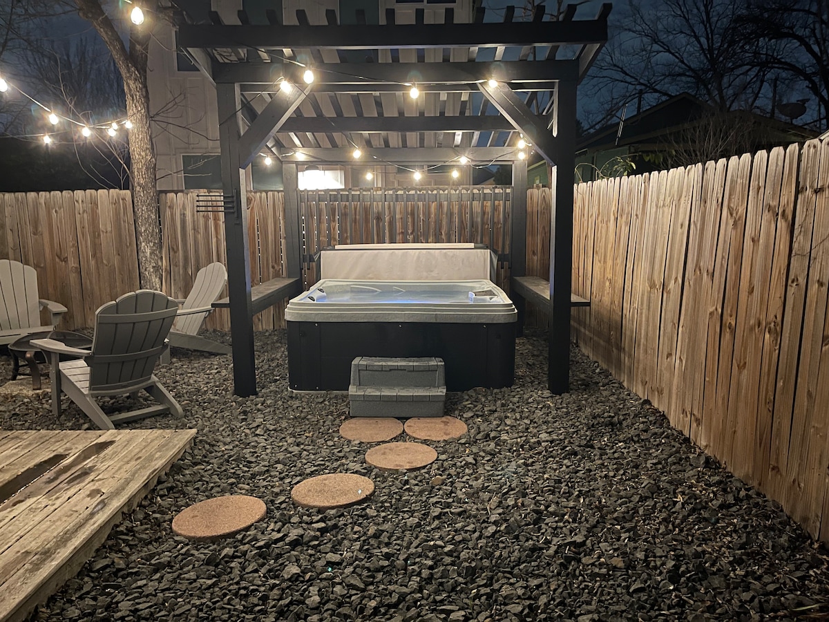 An outdoor area is featured, illuminated by string lights strung overhead. A hot tub is positioned beneath a shaded structure, surrounded by gravel. Several adirondack chairs are arranged nearby, and a pathway of round stepping stones leads to the seating area.