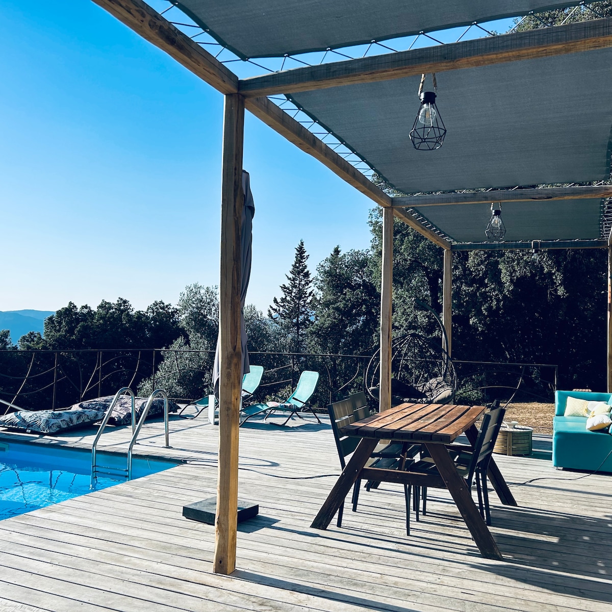 An outdoor deck features a wooden dining table surrounded by chairs, with a swimming pool visible nearby. Sun loungers are positioned to one side, and a shaded area is supported by wooden beams. A clear sky and distant trees can be seen in the background.