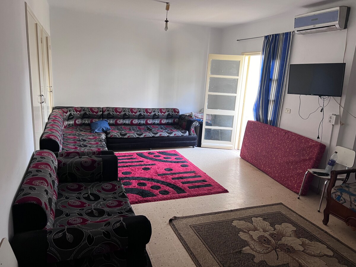 A spacious living area is furnished with a large sectional sofa featuring vibrant patterns. A television is mounted on the wall opposite the seating. Natural light enters through a glass door, enhancing the room's bright ambiance. A patterned area rug complements the overall decor.